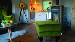 Un espace de vie compact comprenant un fauteuil vert, une petite table avec des fruits, un lampadaire sur trépied, un coin cuisine et une cheminée ornée d’une statue.