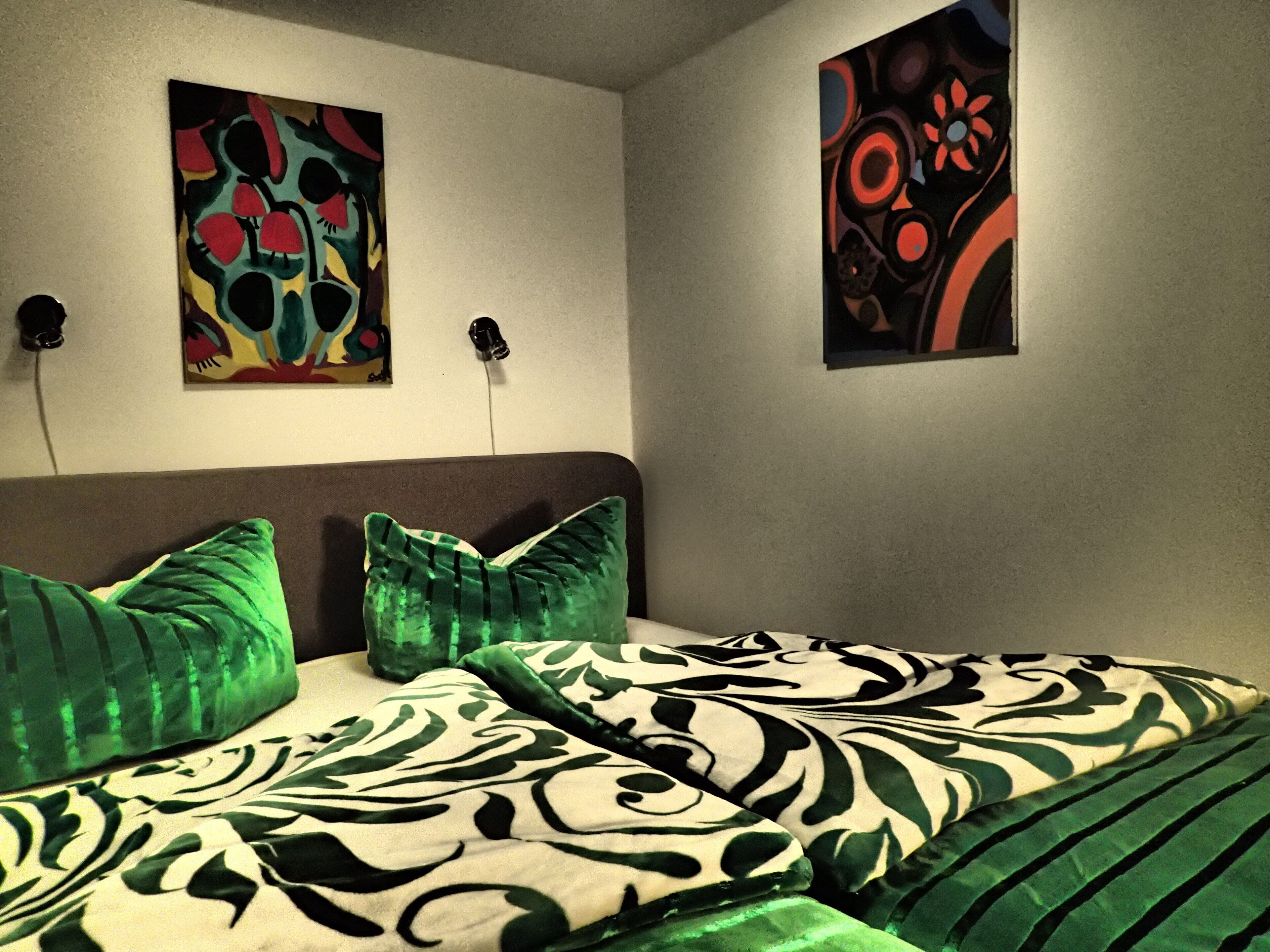Une chambre à coucher avec un lit, des oreillers verts et deux tableaux abstraits accrochés au mur.