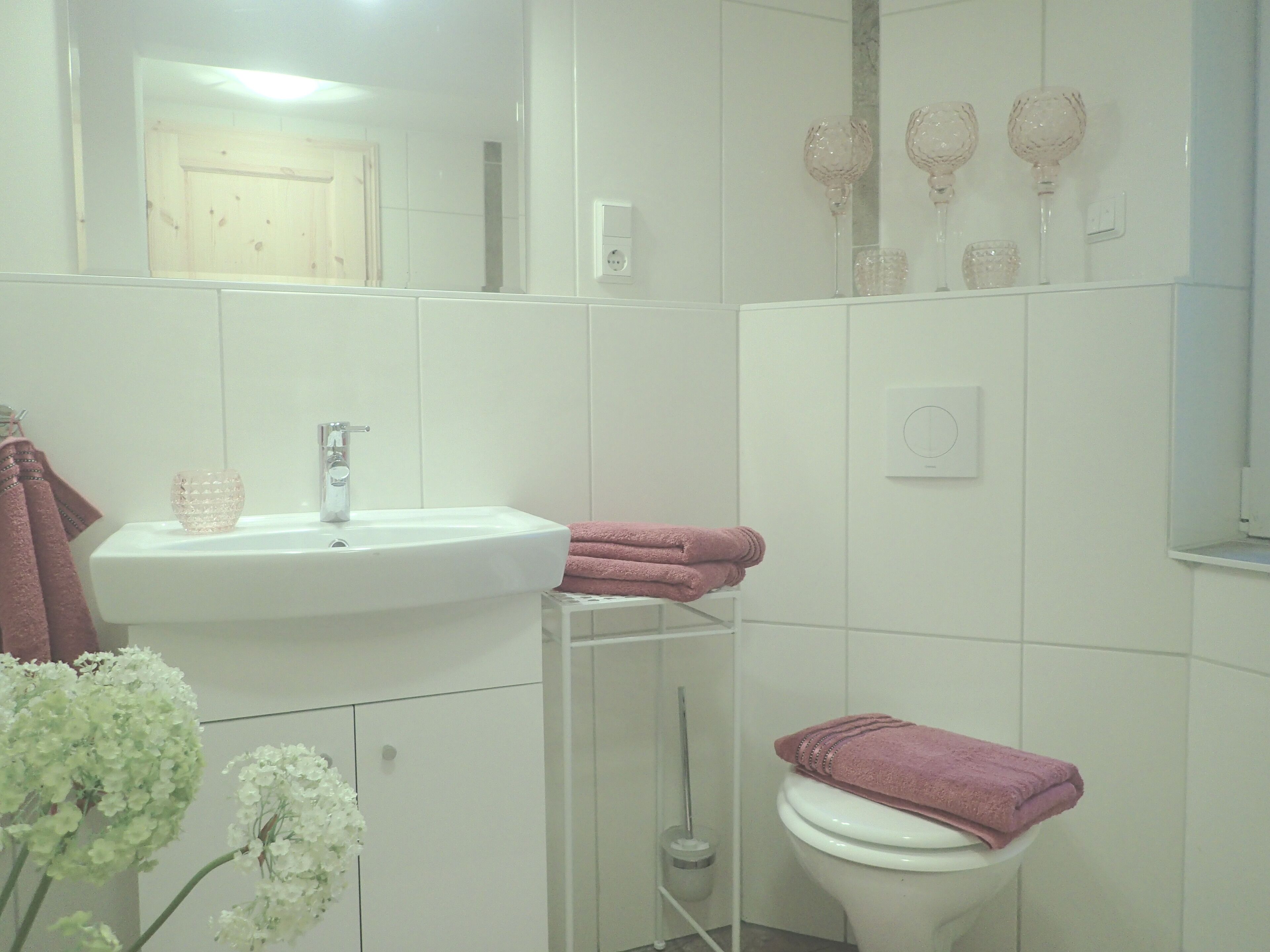 A bathroom with a white sink, a toilet, and a mirror. There are pink towels on the toilet and a glass vase on the sink.