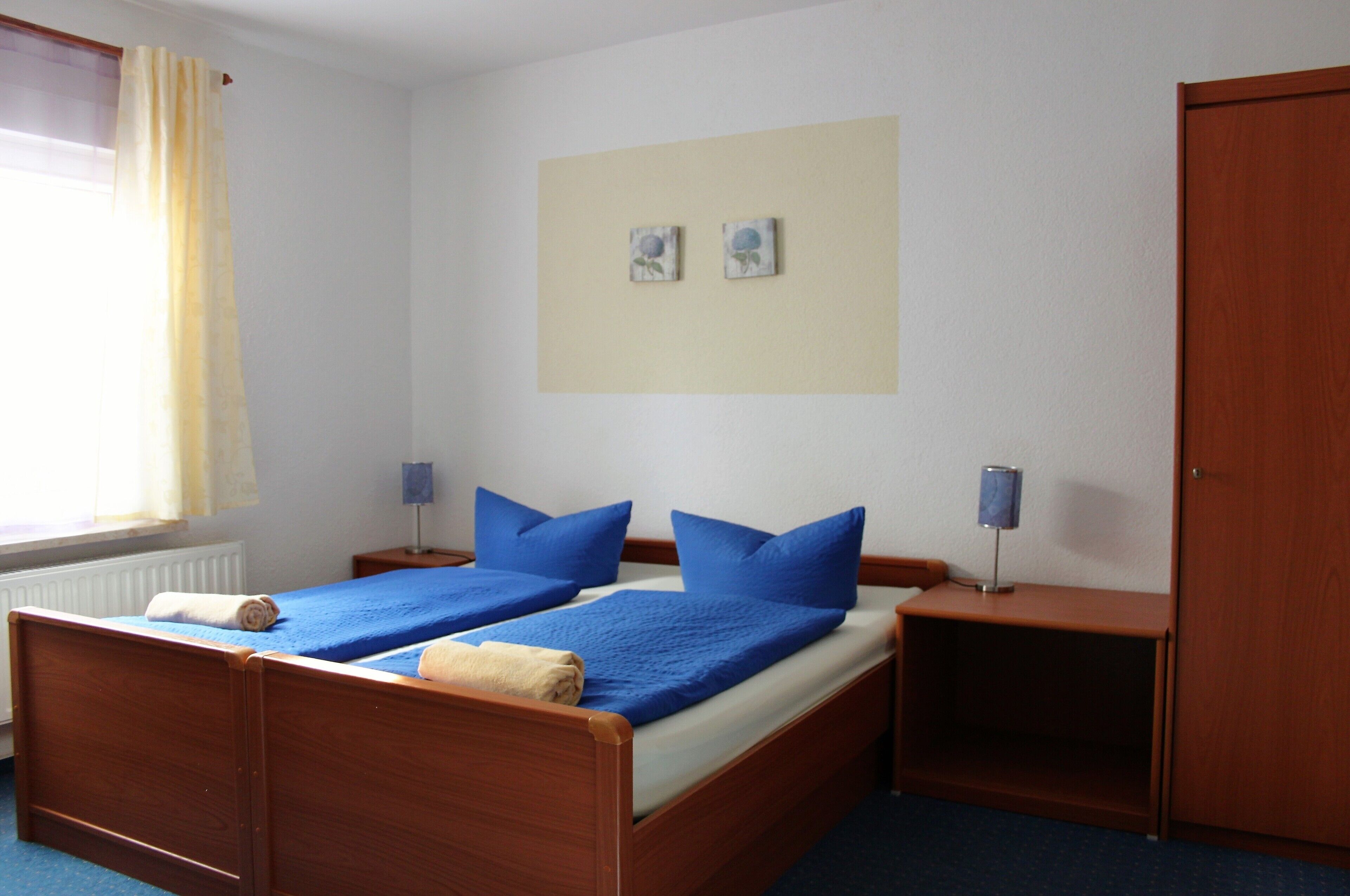 A double bed with blue bedding, two bedside lamps, a wooden wardrobe, and a window with curtains.