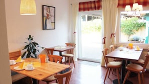A dining area with wooden tables and chairs, a hanging light fixture, a framed picture on the wall, and a window with curtains. - Pension An der Kamske (Lübbenau/Spreewald)