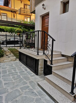 A stone-paved pathway leading to a building with a wooden door and a black metal railing. - Cozy house near the gondola in Bansko.  (Bansko)
