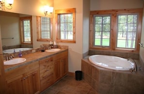 Ein Badezimmer mit zwei Waschbecken, einer Badewanne, Holzschränken und großen Fenstern mit Blick auf Bäume.