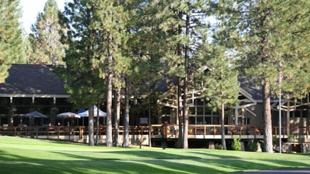 A lodge with a covered deck, surrounded by tall pine trees, and a well-maintained lawn.