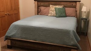 Un dormitorio con estructura de cama de madera, una colcha azul claro y dos almohadas. Hay una mesita de noche con una lámpara y un cuadro pequeño en la pared sobre la cama.