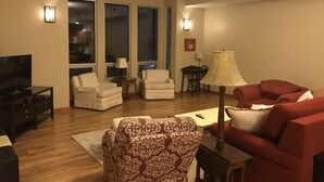 Una sala de estar con televisor, dos sillones blancos, un sillón estampado, un sofá rojo y una mesa de comedor con sillas.