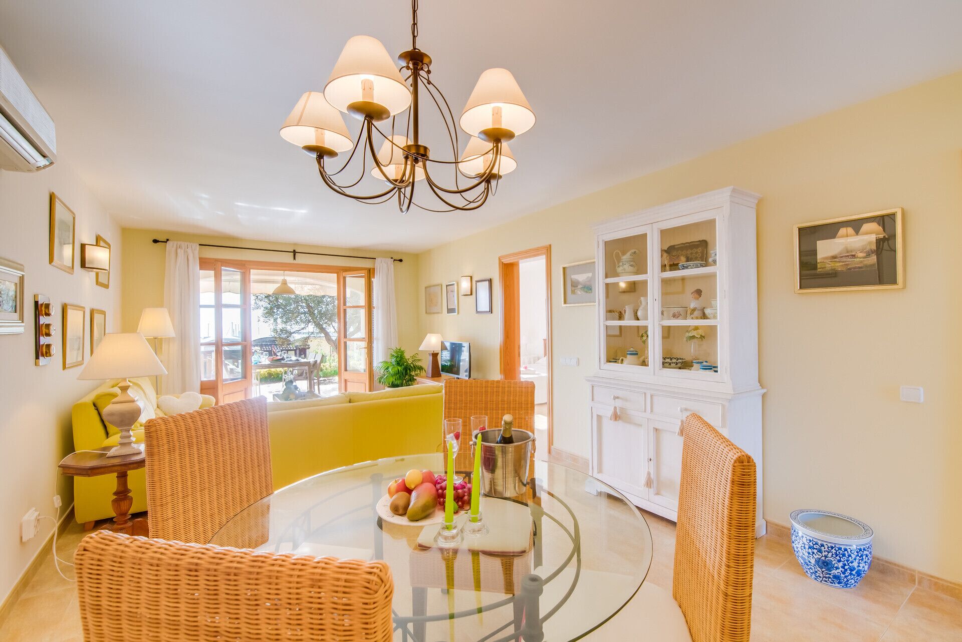 A bright living room with a glass-top table, wicker chairs, a chandelier, and a white cabinet with glass doors.
