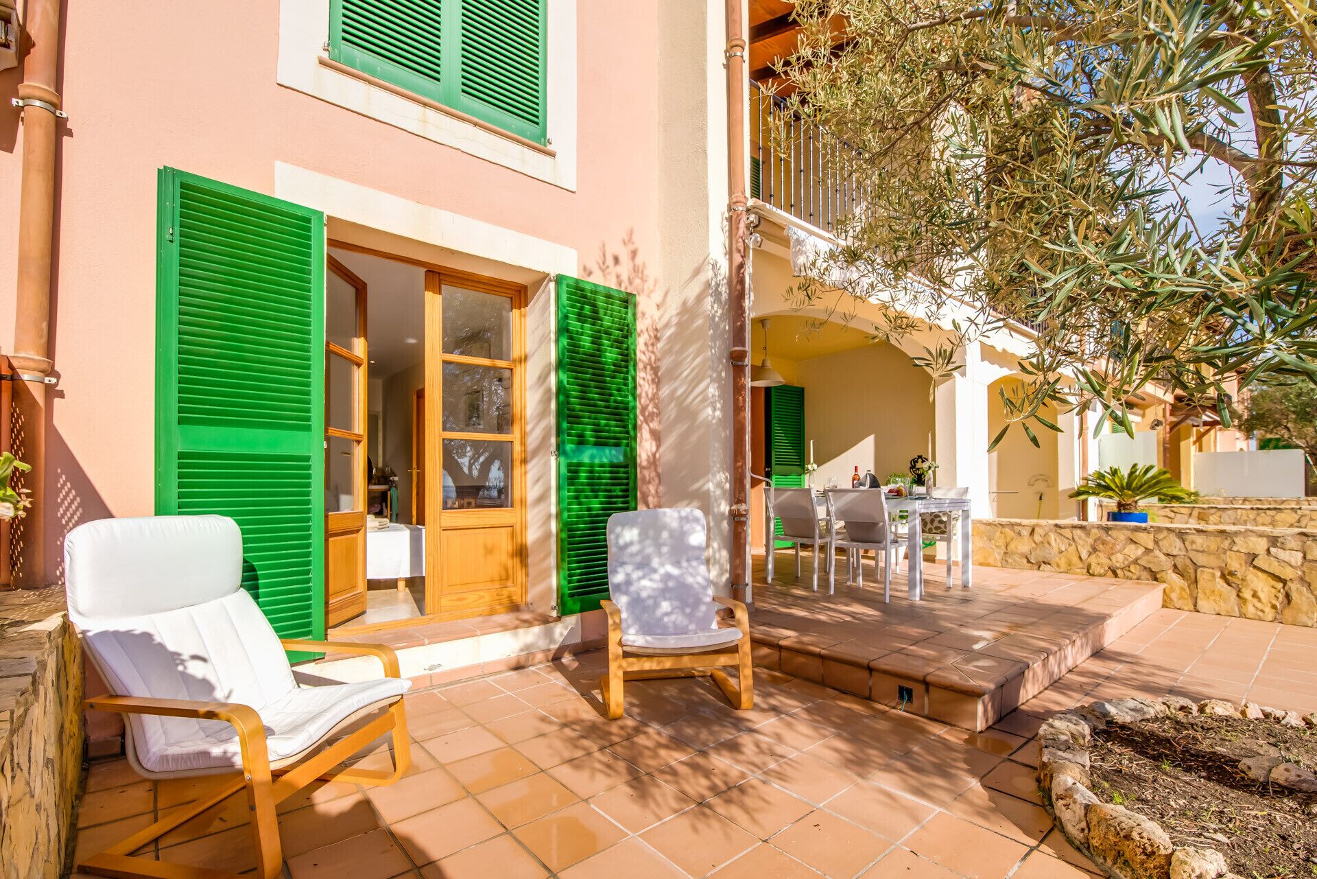 Une terrasse ensoleillée, aménagée à l’extérieur, avec une chaise longue blanche, une chaise en bois et une table entourée de chaises.