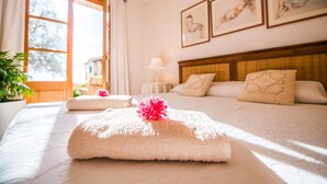 Ein ordentlich bezogenes Bett mit Handtuch und einer rosa Blume, ein Nachttisch mit Lampe und gerahmte Bilder an der Wand.