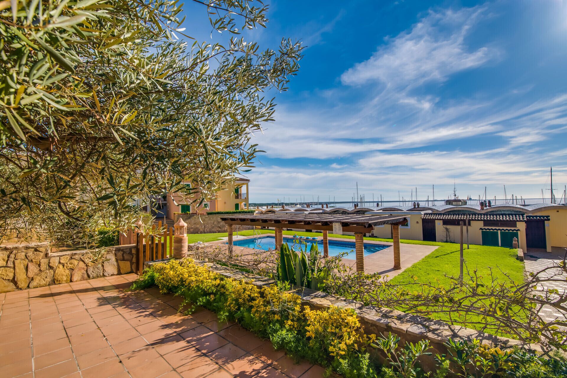 A pool area with a pergola, a stone wall, and a view of the sea with boats.