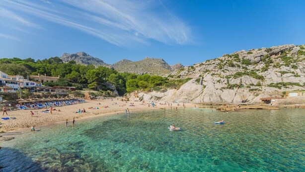 Ein Strand mit klarem Wasser, badenden Personen und Gebäuden im Hintergrund.