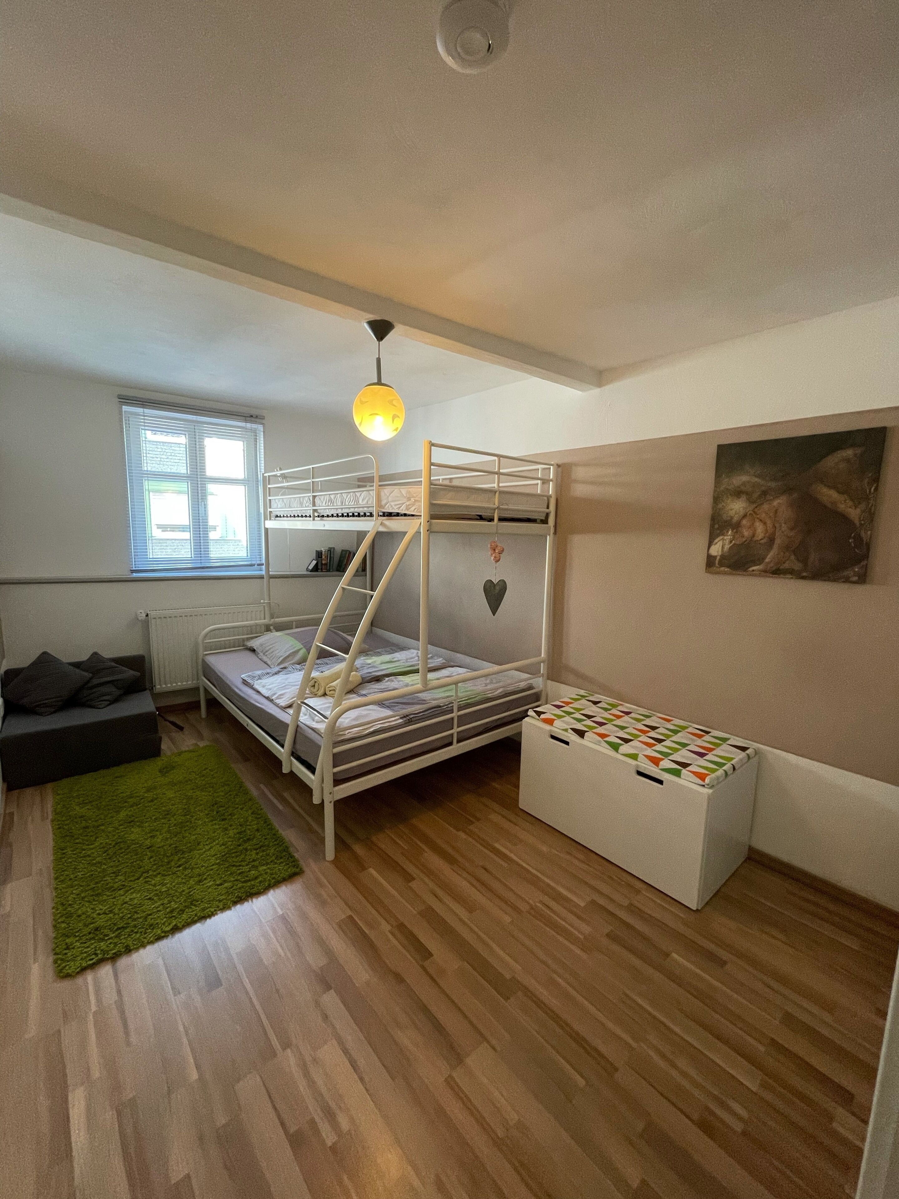 Una habitación pequeña con literas, un sofá compacto, un radiador, una ventana con persianas y un cuadro en la pared.