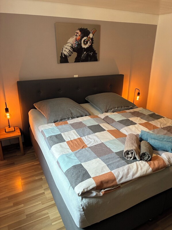 A bed with a checkered blanket, two pillows, and rolled towels. There is a nightstand with a lamp, a wall-mounted artwork, and a wooden floor.