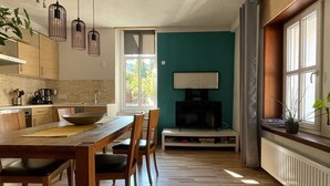 A modern kitchen with wooden cabinetry, a dining table with chairs, pendant lights, a large window with curtains, and a television on a stand.