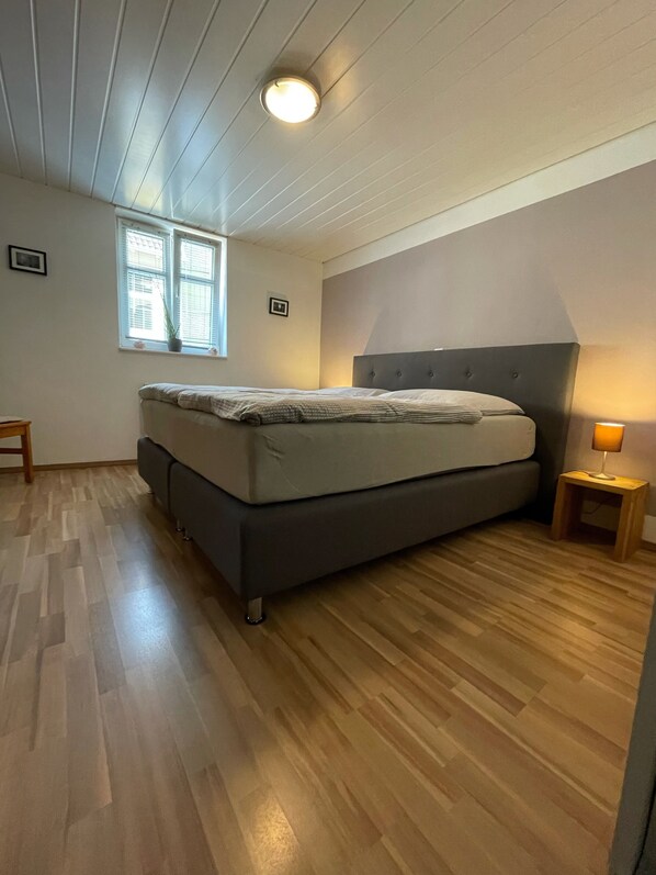 Un dormitorio con una cama, una mesita de noche con lámpara, una ventana con persianas y un taburete pequeño.