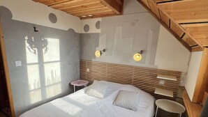 Een slaapkamer met een schuine plafond, houten balken, een groot bed met wit beddengoed, twee nachtkastjes en een wandlamp.