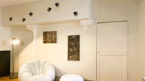 Una habitación con una puerta blanca, dos sillones tipo pouf blancos, una lámpara de pie y tres esculturas de aves fijadas a la pared.