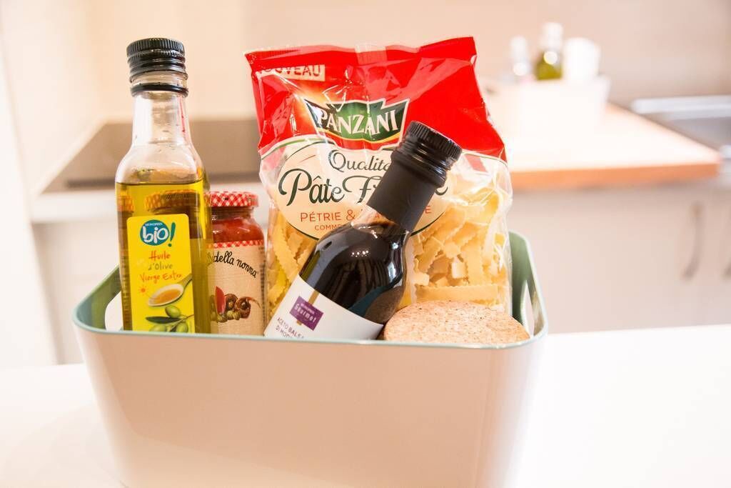 A kitchen counter with a bottle of olive oil, a bag of Panzani pasta, a bottle of wine, and a jar of jam.