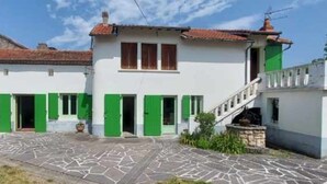 Een huis met twee verdiepingen, voorzien van dakpannen, groene luiken en een erf met stenen bestrating.