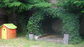 Een houten poppenhuisje met een groen dak en een rode deur, gelegen in een tuin met een stenen muur en weelderige begroeiing op de achtergrond.