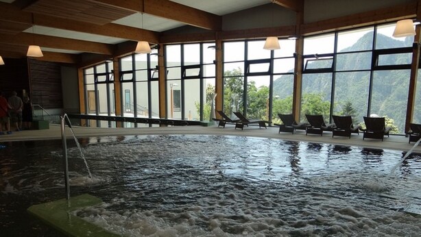 An indoor swimming pool with a whirlpool feature, surrounded by lounge chairs and large windows offering a view of the outdoors. - Gites de Mon Clocher 2 apartments in a village house (Alpes-Maritimes)