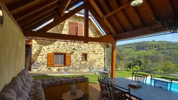 A covered outdoor area with a wooden deck, a stone building with shutters, a pool, and a view of green hills. - Former sheepfold renovated with beautiful view of the Pyrenees ideal for the family (Astugue)