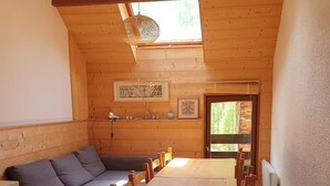 Ein gemütliches Zimmer mit Holzbalkendecke, einer Couch, einem Esstisch mit Stühlen und einem Fenster mit Blick ins Grüne.