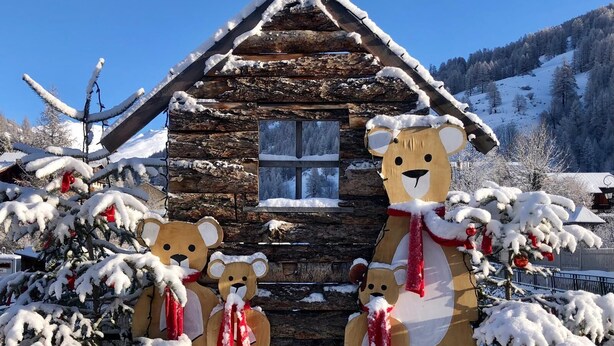 Eine Holzhütte mit schneebedecktem Dach und Fenster, umgeben von schneebedeckten Bäumen und blauem Himmel.