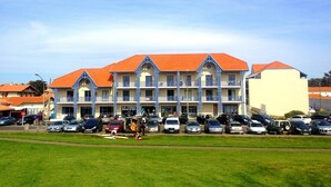 A multi-story hotel with a red roof and blue balconies. There are several cars parked in front, and a grassy area in the foreground. - Beautiful, warmly decorated apartment-1st line sea-Pool (Biscarrosse)