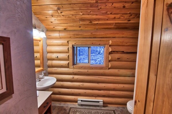 Un baño de cabaña de madera con interior de madera, una ventana que ofrece vistas a árboles cubiertos de nieve, un lavabo y un inodoro.