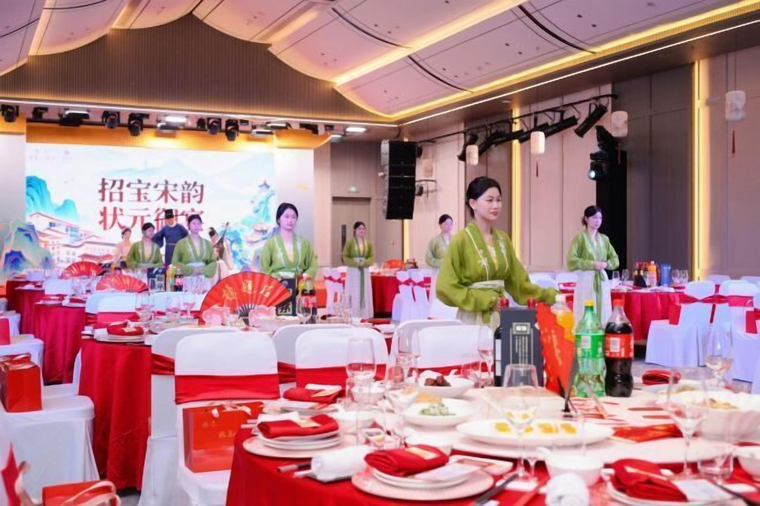 A banquet hall with round tables set for a formal event, featuring red and white tablecloths and a stage with a backdrop.