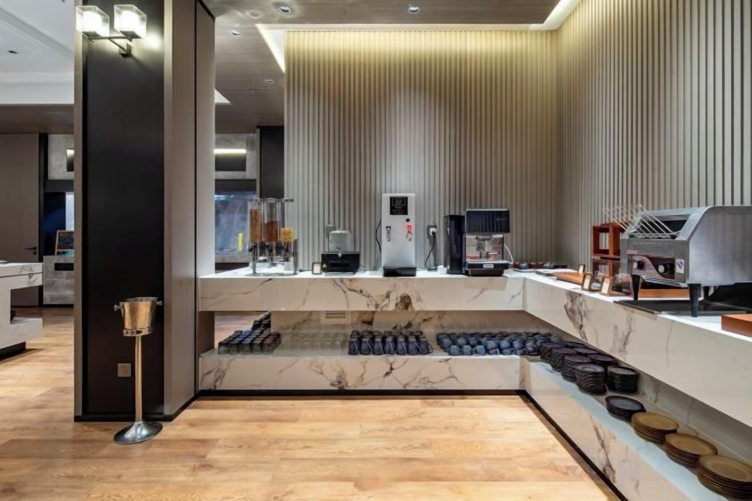 A modern hotel lobby with a marble reception desk, a coffee machine, and a seating area with a wooden floor.