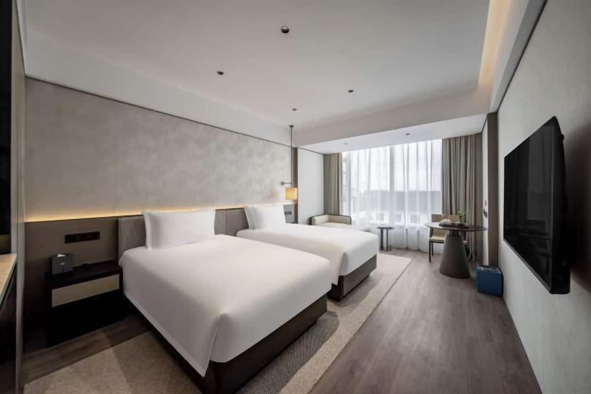 A modern hotel room with two beds, a TV, a desk, and a view of the city.