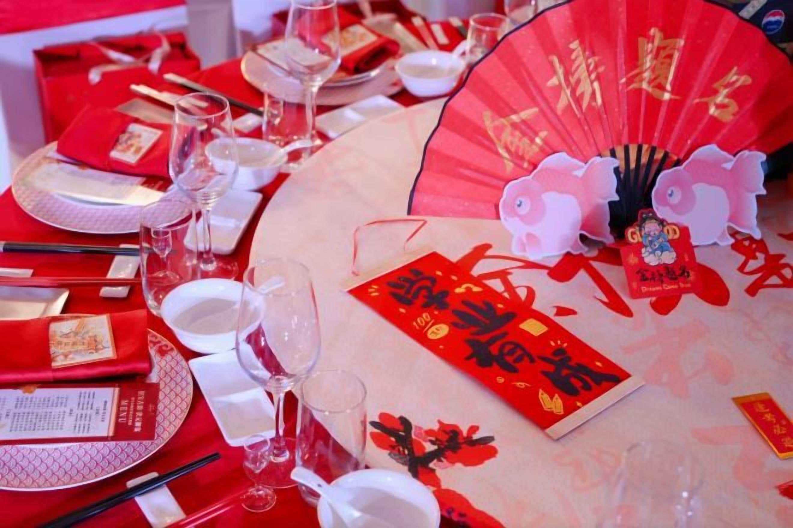 Ein Tisch ist gedeckt mit Geschirr in Rot und Weiß, darunter Teller, Schüsseln und Stäbchen, zusammen mit einem roten Fächer und einem roten Umschlag mit goldener Schrift.