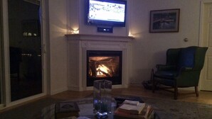 Een woonkamer met een open haard, een televisie erboven en een groene fauteuil.