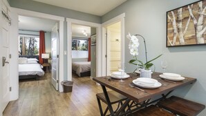 A room with a wooden dining table set for two, a bed with white bedding, and a small trash bin. - French Street Haven | 5 Minute Walk to Main Street (Breckenridge)