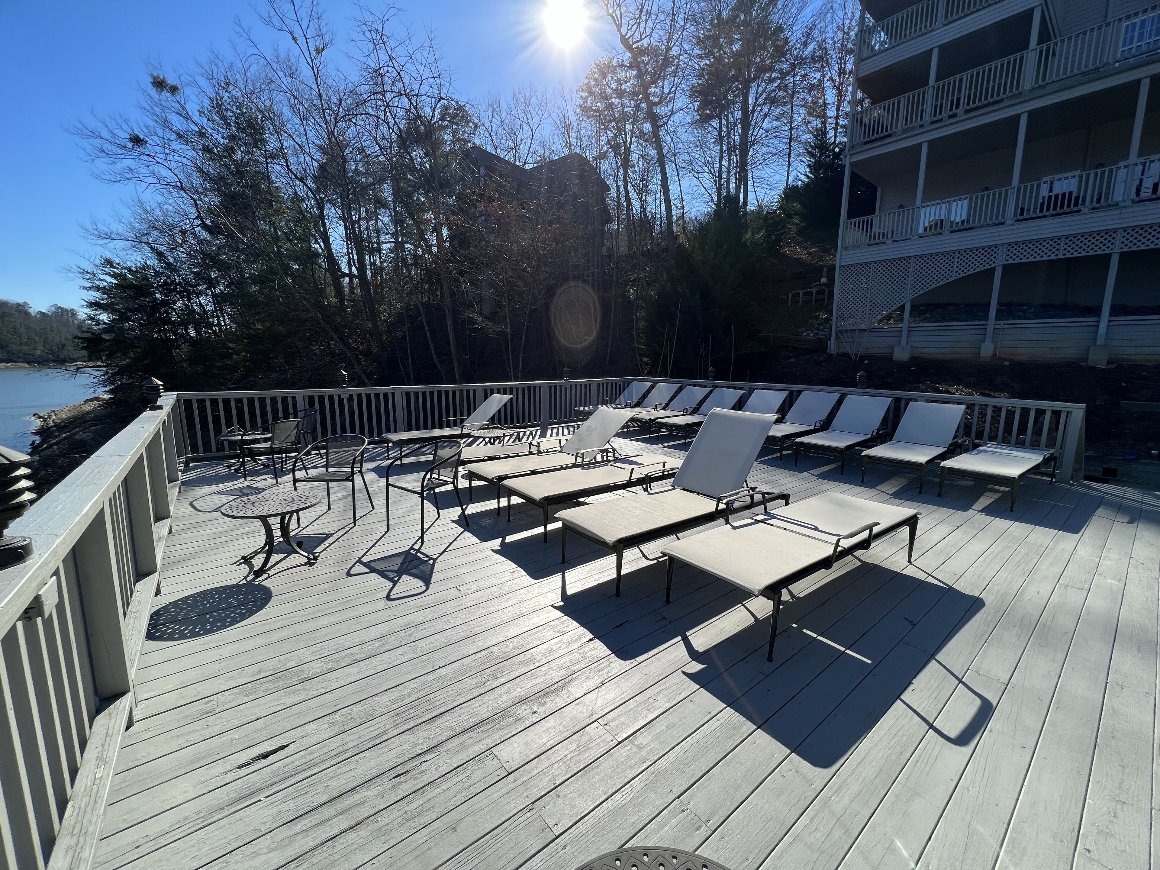 Une terrasse en bois, dotée de chaises longues et de tables, qui offre une vue sur une étendue d’eau et des arbres.