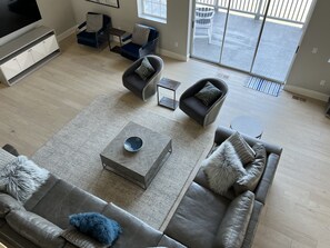 Un salon moderne avec un canapé gris, une table basse et un téléviseur posé sur un meuble. On y trouve deux fauteuils et une petite table. La pièce est dotée d’une grande porte-fenêtre coulissante qui donne sur un espace extérieur.