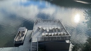 En bro med en båd, et overdækket siddeområde med borde og stole, og udsigt til en rolig sø med bjerge i baggrunden.