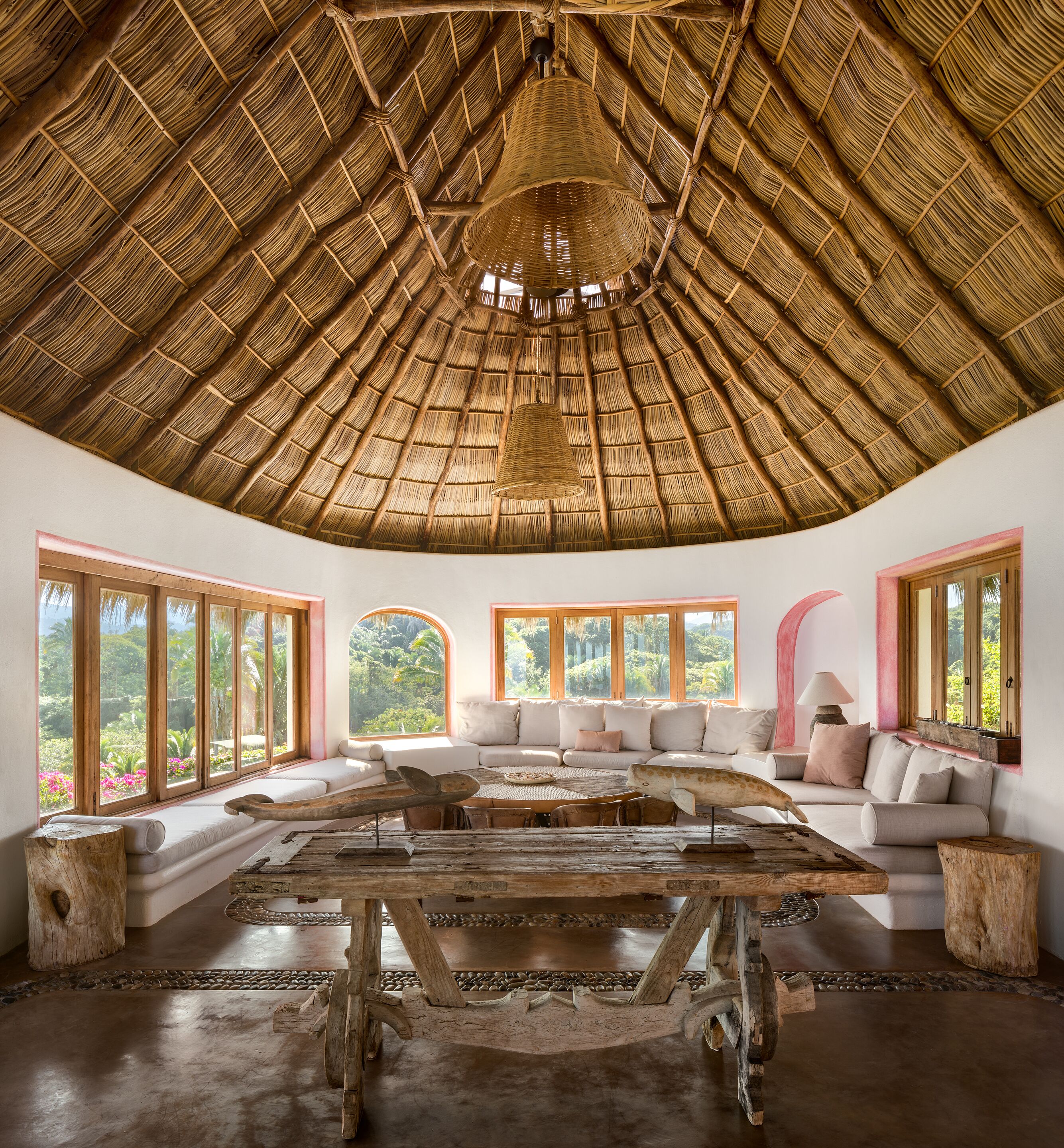 A room with a thatched roof, large windows, a wooden table, and a sofa.