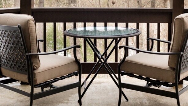 Een overdekt terras met twee stoelen en een tafel, met uitzicht op een bebost gebied.