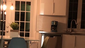 A kitchen with white cabinets, a coffee maker, and a sink. There is a dining table with chairs and a potted plant on the windowsill.