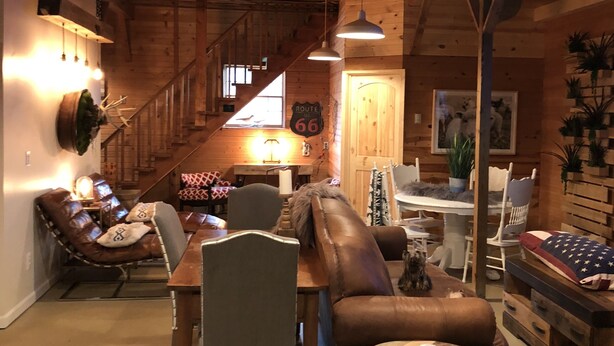 A cozy cabin interior with a wooden ceiling, a dining area with a table and chairs, a leather sofa, and a staircase leading to an upper level.