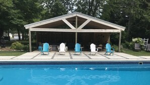 A swimming pool with a covered seating area and Adirondack chairs.