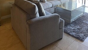 Un salon avec un canapé gris, une table basse en verre et un ventilateur de plafond. Un tableau encadré est accroché au mur, et il y a une fenêtre avec des rideaux.