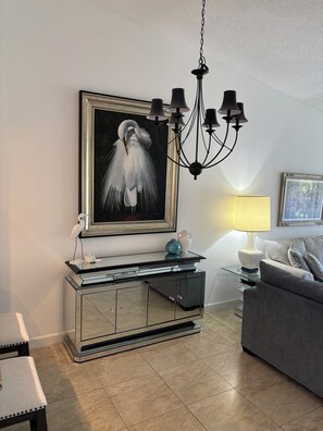 A modern living room with a glass coffee table, a framed painting of a bird, a chandelier, and a grey sofa.