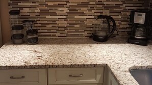 Une cuisine avec des meubles blancs, un dosseret carrelé et un plan de travail en granit. On y trouve deux machines à café et une bouilloire sur le plan de travail.