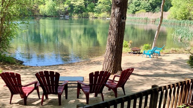 A lakeside seating area with red chairs and a table, surrounded by trees and a wooden fence. - Waterfront Plymouth Pond Retreat, Sleeps 6, Pet-Friendly, Kayaks, Fire Pits (Plymouth)