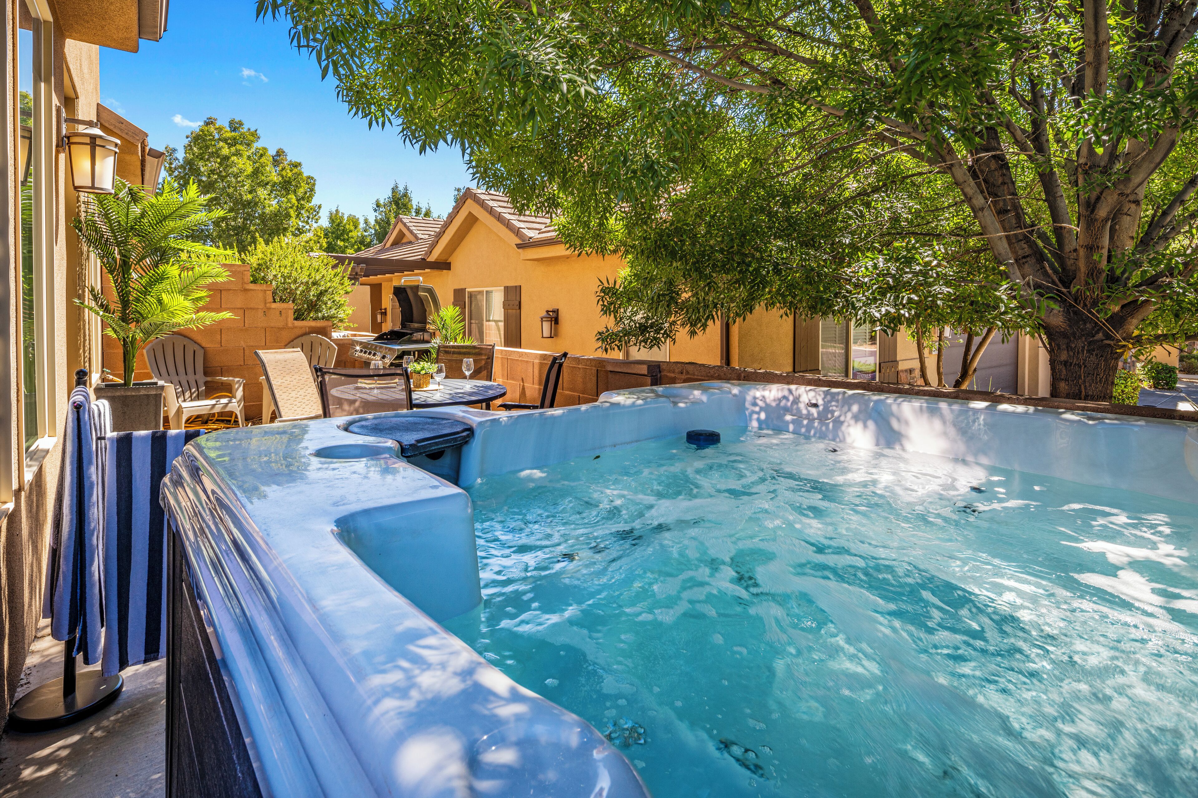 A backyard with a hot tub, patio furniture, and a tree providing shade.
