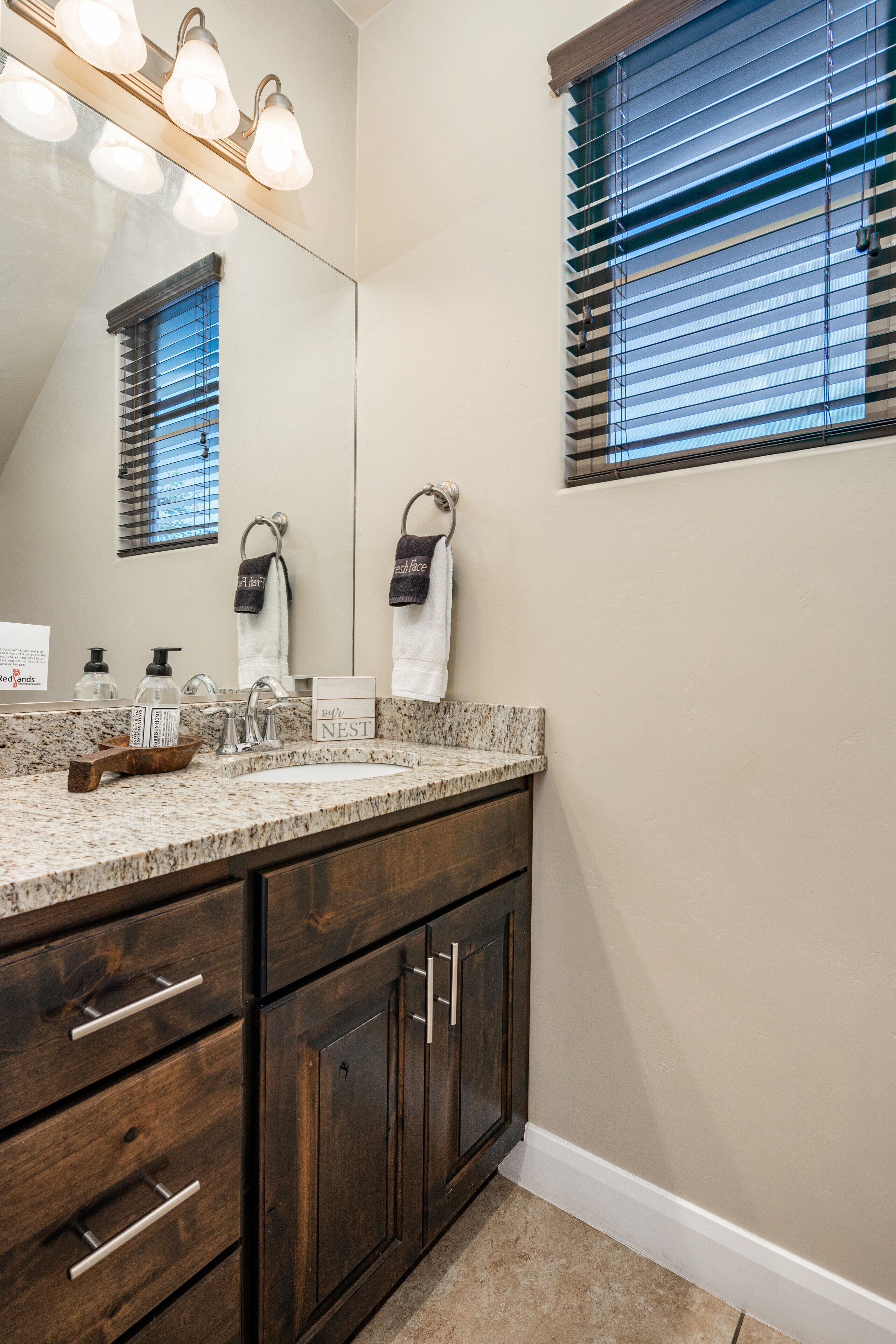 A bathroom with a granite countertop, dark wooden cabinets, a large mirror, and a window with blinds.